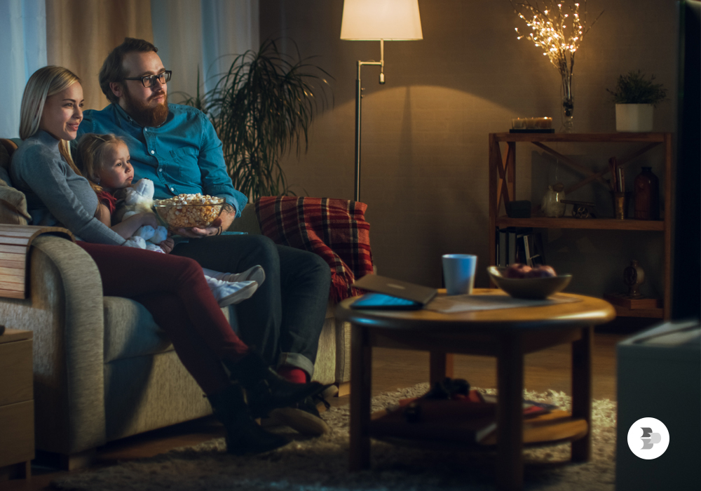 Homem, mulher e menina sentados no sofá comendo pipoca e assistindo televisão.