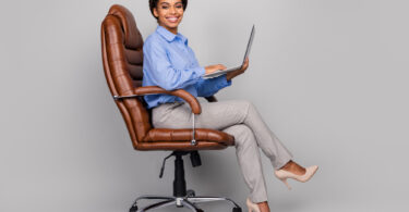 Uma moca usando seu notebook em cima de uma cadeira de escritório. Cadeira escritório.