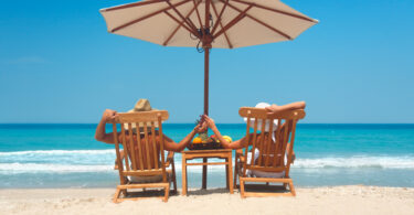 Um casal sentado nas cadeiras praia, com um guarda-sol em cima. Guarda-sol.