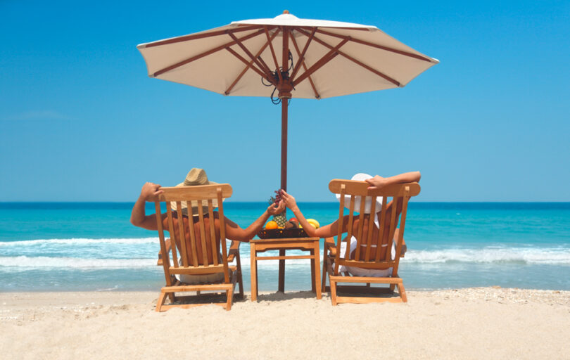 Um casal sentado nas cadeiras praia, com um guarda-sol em cima. Guarda-sol.