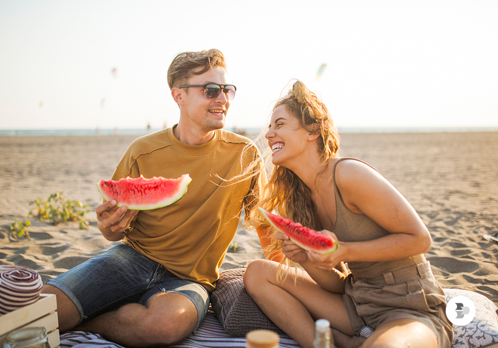 Um casal jovem comendo melancia na beira da praia. Frutas de verão.